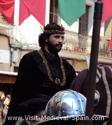 In this photo, King Pere III arrives in Manresa on horesback escorted by members of his court and knights in armour.There are coloured flags and banners hanging from balconies in the background.